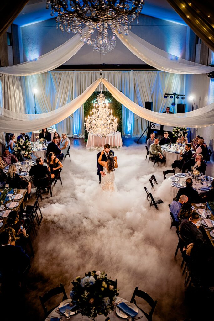 Bride and groom first dance surrounded by dance on the clouds fog effect in the Ranch House ballroom at Bakers Ranch wedding venue in Parrish FL with three crystal chandeliers and white draped ceiling