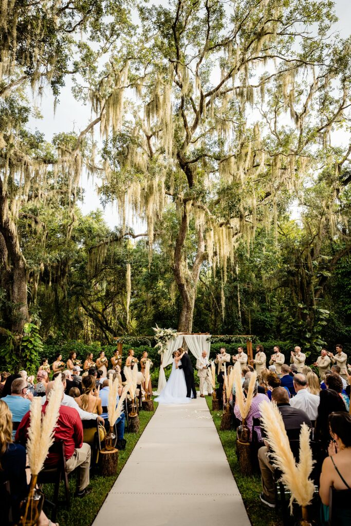 Outdoor wedding ceremony at Ancient Oaks ceremony site at Bakers Ranch wedding venue in Parrish FL with massive live oak trees draped in Spanish moss pampas grass aisle decor and full guest attendance