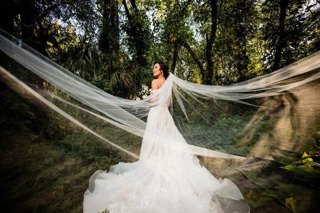Asian bride with cathedral veil spread wide in the hidden wooded area at Bakers Ranch wedding venue in Parrish FL with lush Florida forest in the background