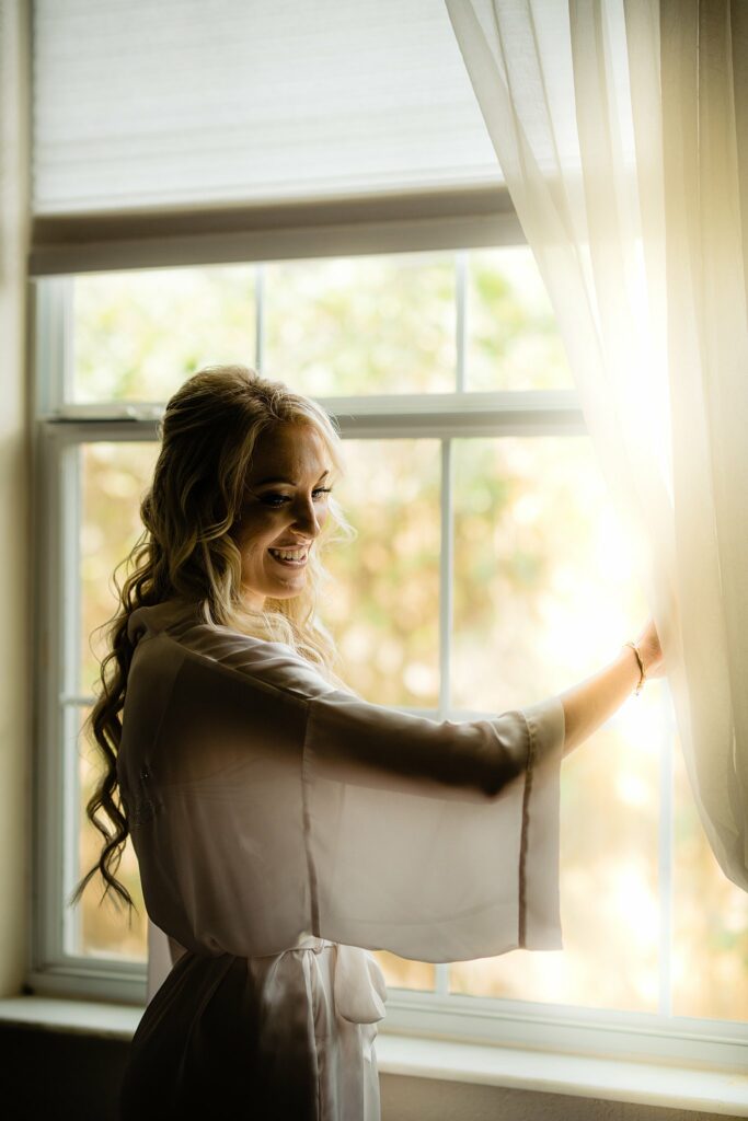 Bride in robe looking out the window of the bridal suite at Bakers Ranch wedding venue in Parrish FL with natural window light