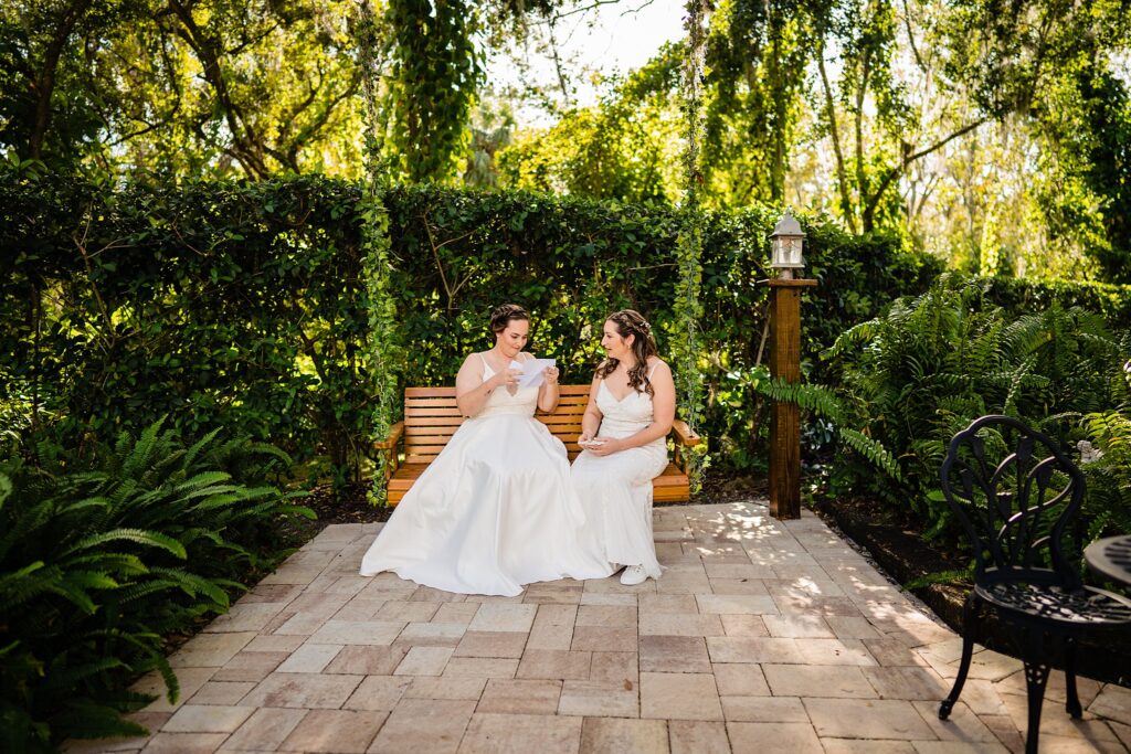Two brides reading vow books together on the wooden garden swing at Bakers Ranch wedding venue in Parrish FL surrounded by lush greenery and ferns