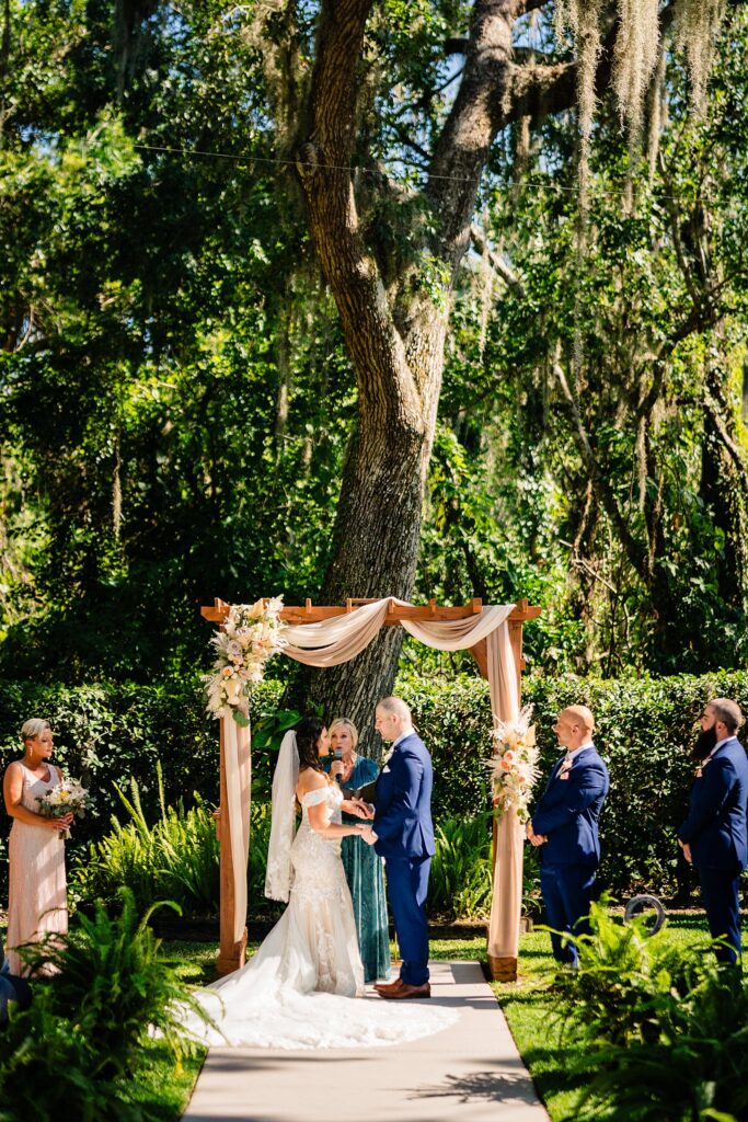 Outdoor wedding ceremony at Ancient Oaks ceremony site at Bakers Ranch wedding venue in Parrish FL with bride and groom at wooden arch surrounded by mature oaks and Spanish moss