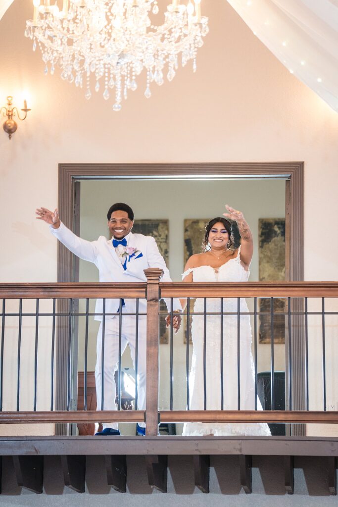 Bride and groom waving from the second floor balcony overlooking the ballroom at Bakers Ranch wedding venue in Parrish FL with chandelier above