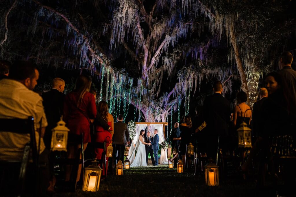Night wedding ceremony at Love Lock Bridge at Bakers Ranch in Parrish FL with massive oak tree lit in purple and teal with fairy lights cascading through Spanish moss