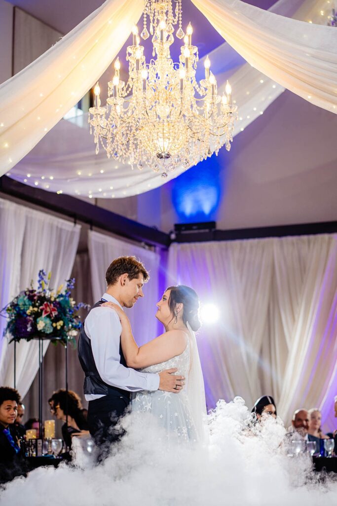 Bride and groom slow dancing surrounded by dance on the clouds fog effect under lowered crystal chandelier in the Ranch House ballroom at Bakers Ranch wedding venue in Parrish FL