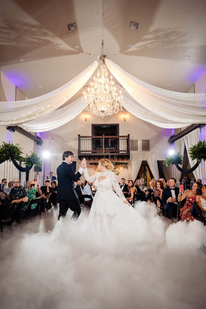 Bride and groom dancing on the clouds fog effect during first dance in the Ranch House ballroom at Bakers Ranch wedding venue in Parrish FL with crystal chandelier and white draped ceiling and second floor balcony visible in background