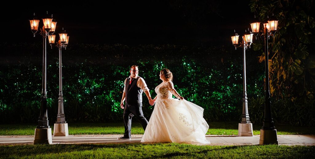 Bride and groom walking hand in hand on the gas lamp pathway between Enchanted Garden Chapel and Infinity Forest at Bakers Ranch wedding venue in Parrish FL at night with green uplighting on hedges