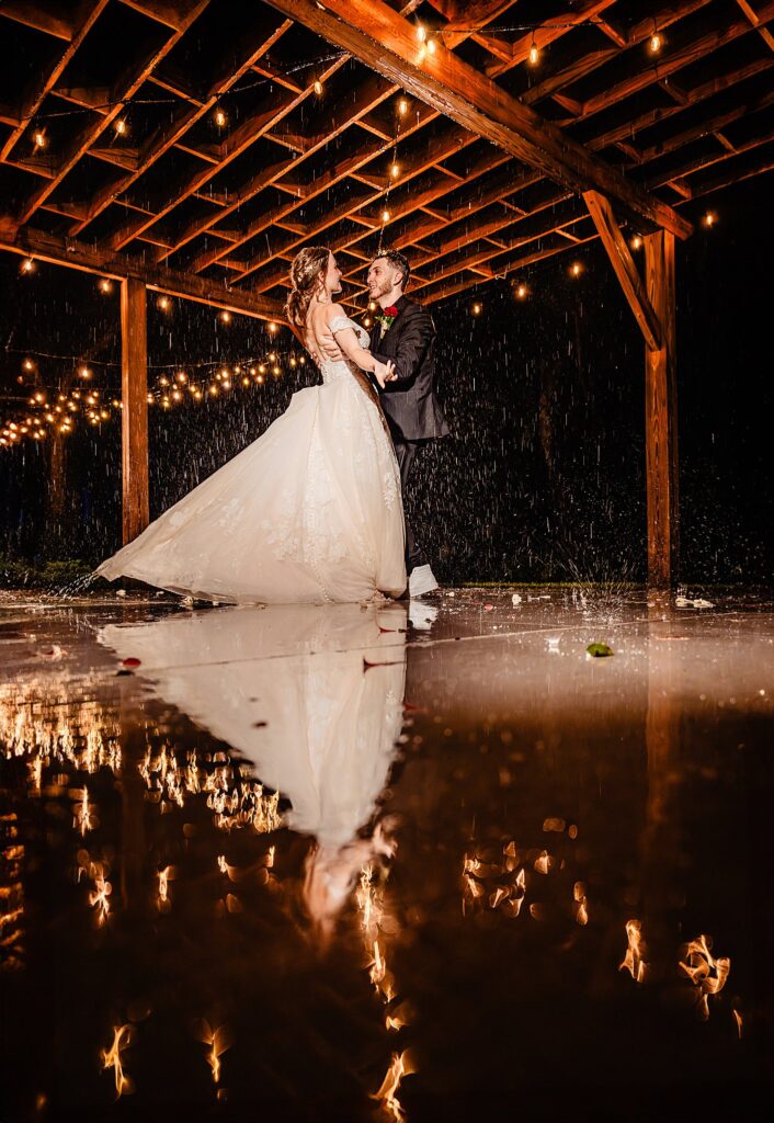 Bride and groom dancing in the rain under the timber pergola gondola area at Bakers Ranch wedding venue in Parrish FL with Edison bulb string lights and reflection on flooded patio stones