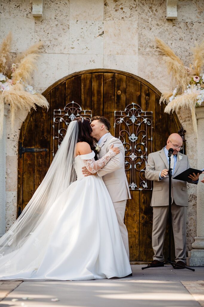 Bride and groom sharing first kiss in front of the arched wooden doors with wrought iron ironwork at Enchanted Garden Chapel at Bakers Ranch wedding venue in Parrish FL with pampas grass florals