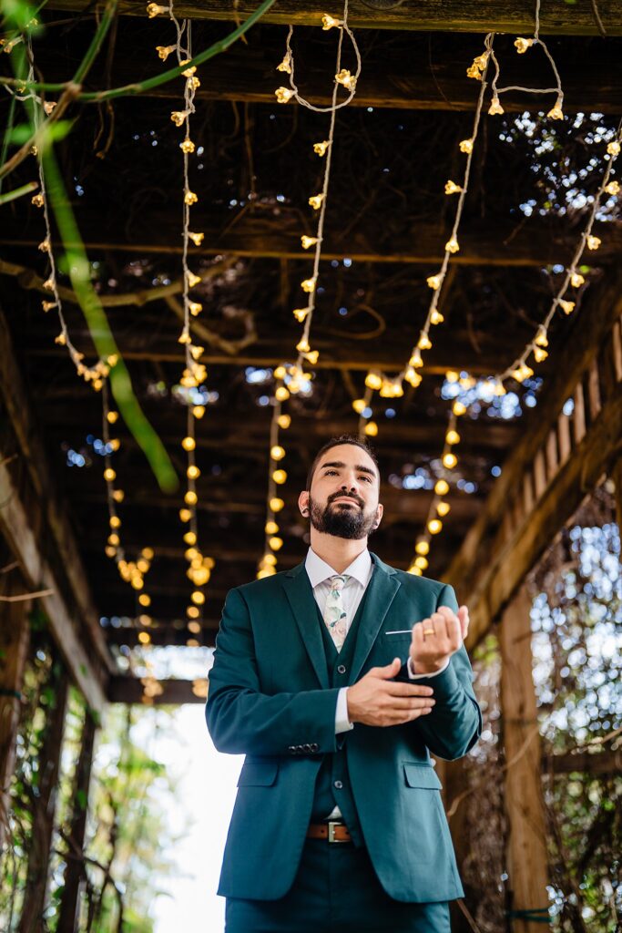 Groom in teal suit adjusting cufflinks beneath string lights at the Tunnel of Love at Mixon Farms, Bradenton Florida