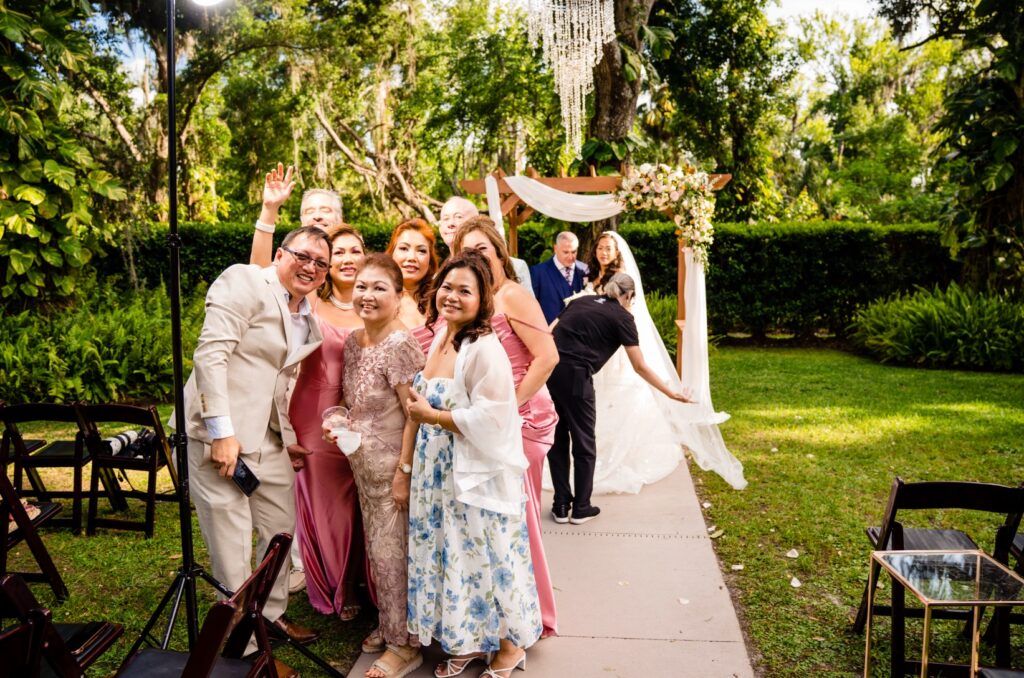 Extended family posing for an impromptu group photo directly in front of the photographer while she adjusts the bride's cathedral veil for family formals at an outdoor wedding ceremony arch