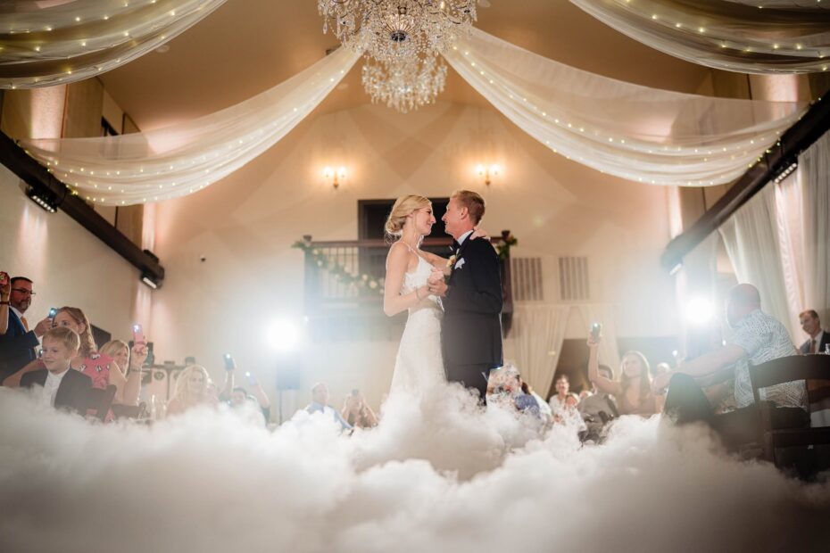Bride and groom sharing their first dance surrounded by a low cloud effect beneath a crystal chandelier and draped ceiling inside the Tudor ranch house at Bakers Ranch