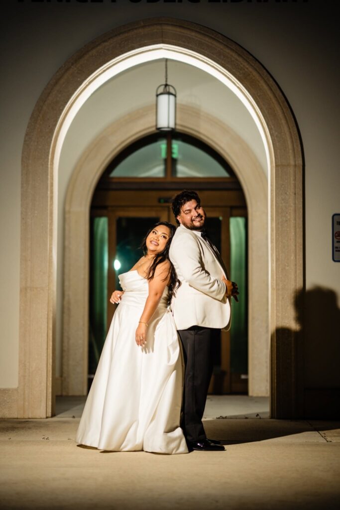 Bride and groom standing back to back under the illuminated stone arch at the Venice Public Library at night after their Filipino-Mexican wedding, photographed by Ying Multimedia