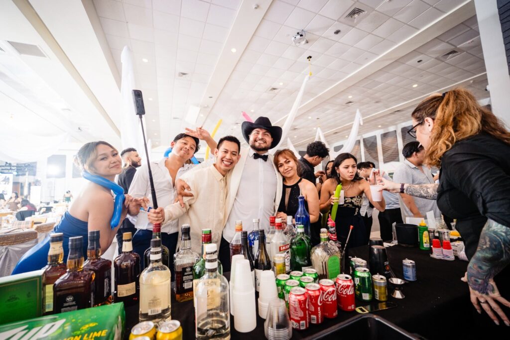 Groom in white tuxedo and cowboy hat posing with guests and a selfie stick at the bar during his reception at the Venice Community Center, Florida wedding photographed by Ying Multimedia