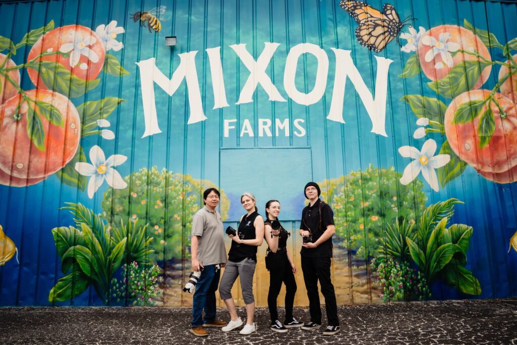 The Ying Multimedia photography team standing in front of the Mixon Farms mural with cameras at the grand reopening