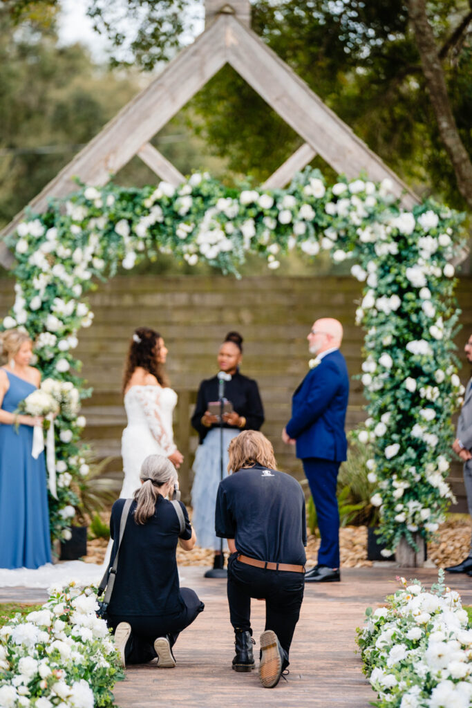 Photographer Rachel and cinematographer Jo kneeling side by side at the base of the aisle capturing a wedding ceremony under a white floral arch